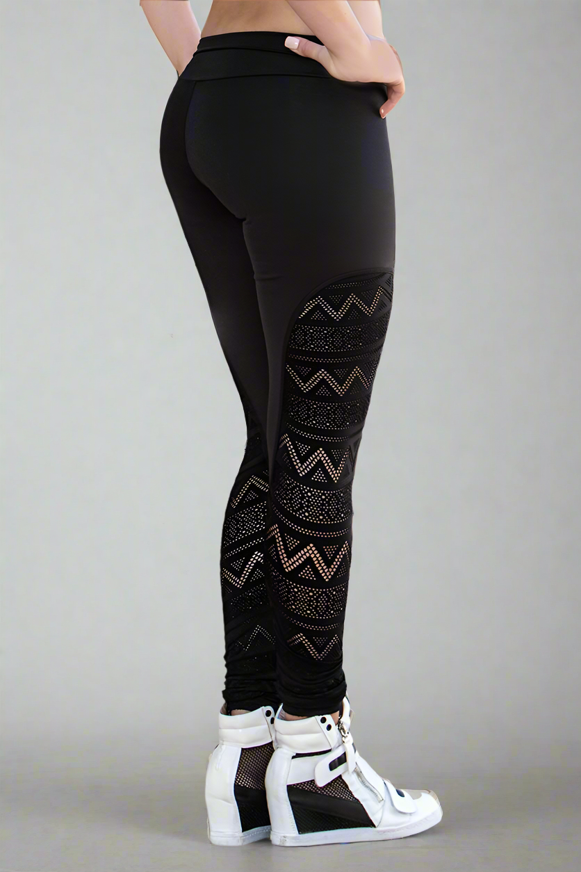A pair of black high-waisted leggings with geometric crochet lace detailing at the ankles and contrasting color accents, worn with white sneakers.